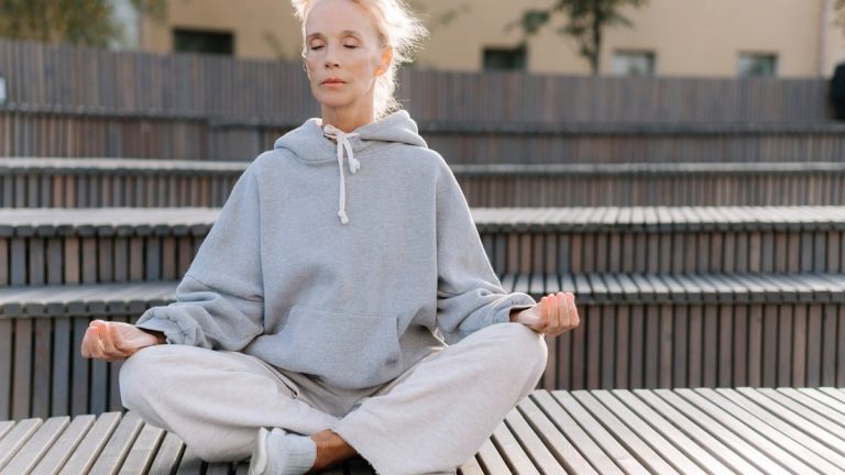 Elderly woman in gray hoodie meditating outdoors, promoting fitness and relaxation.